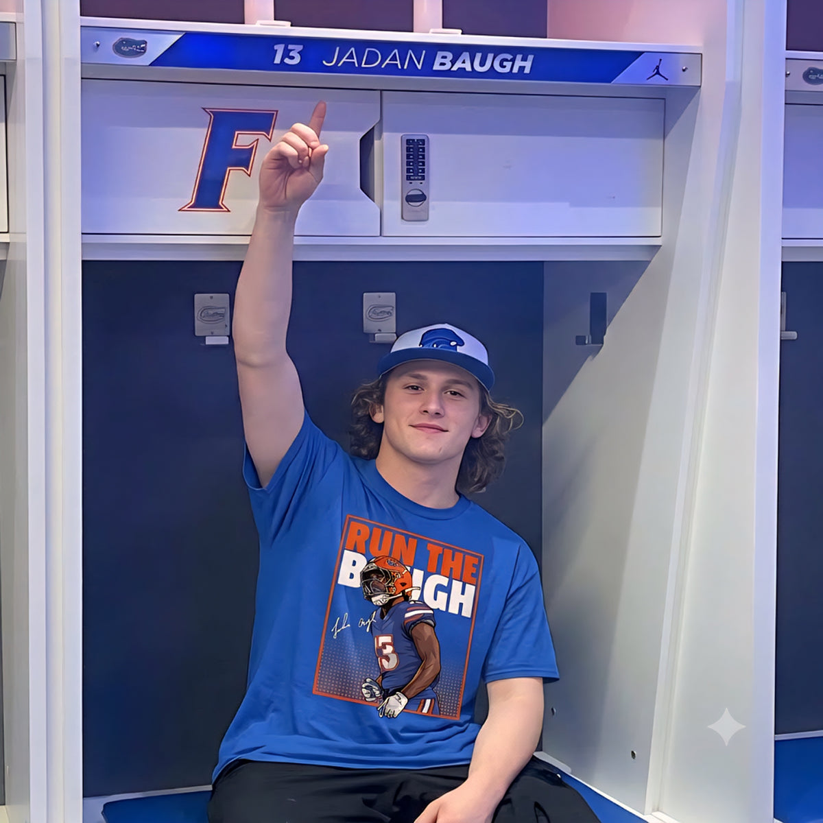 A young man sitting in a locker wearing a blue graphic T-shirt that reads 'RUN THE BOUGH' and a matching blue cap, with his finger pointing upward toward the locker shelf labeled '13 JADAN BAUGH'.