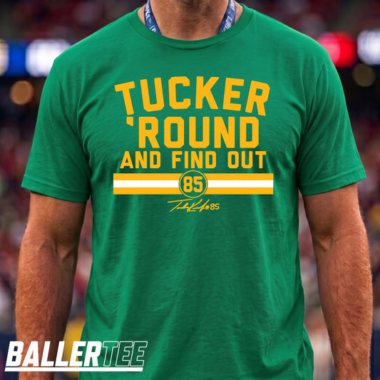 A man wearing a green short-sleeve graphic T-shirt with yellow and white text reading 'TUCKER ROUND AND FIND OUT' and '85' on the front. The photo is cropped from the neck to the waist, with a blurred stadium-like background and a 'BALLERTEE' watermark at the bottom left.