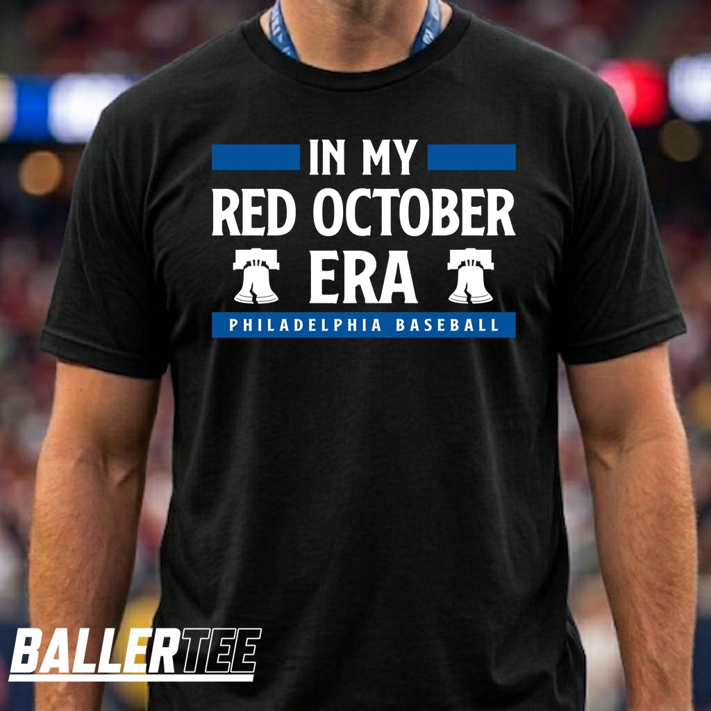 A man wearing a black short-sleeve t-shirt with a blue and white graphic that reads 'IN MY RED OCTOBER ERA PHILADELPHIA BASEBALL' with two Liberty Bell icons. The BallerTee logo is visible in the bottom-left corner.