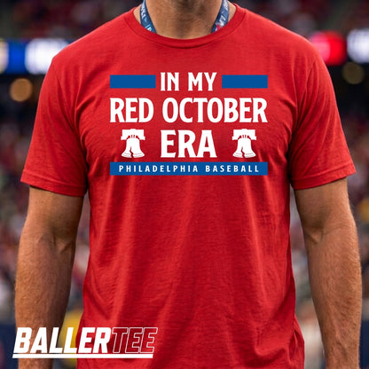 A man wearing a red graphic t-shirt with white and blue text reading 'IN MY RED OCTOBER ERA PHILADELPHIA BASEBALL' and two white Liberty Bell graphics. The BallerTee logo is visible in the bottom-left corner.