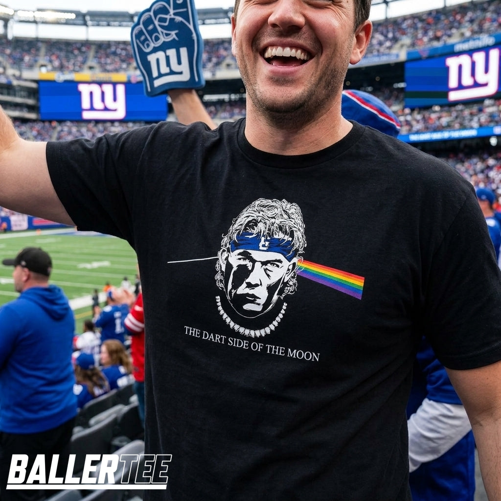 A man wearing a black BallerTee t-shirt with a graphic of a face, a blue headband with a 'B', and a rainbow stripe, standing in a stadium. The shirt text reads 'THE DART SIDE OF THE MOON'.