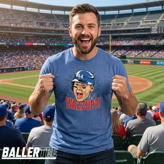 A man in a blue stadium stands smiling with fists raised, wearing a blue T-shirt with a cartoon caricature of himself wearing a blue cap and the text 'WAZZUP!!' in red. A baseball field and crowded stands are visible in the background.