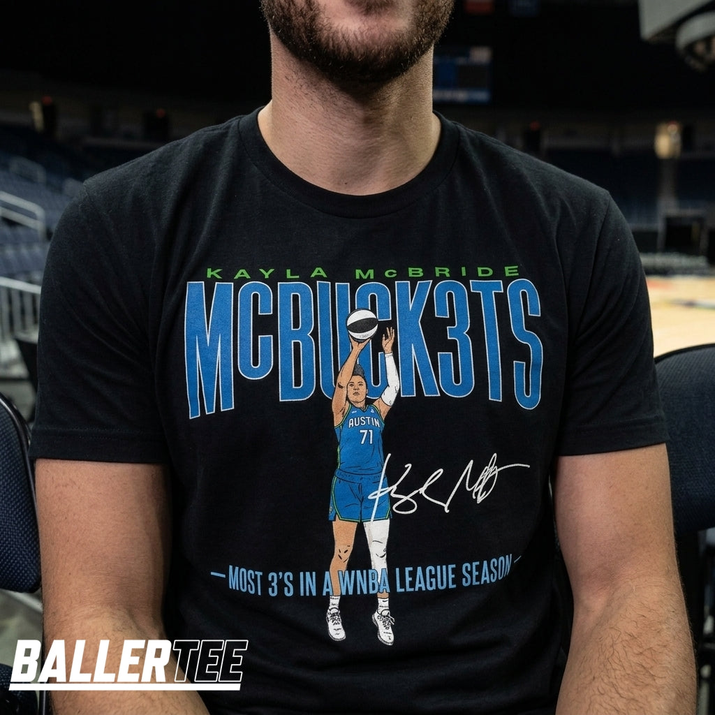 A man wearing a black graphic t-shirt featuring a blue and green design that reads 'KAYLA MCBRIDE MCBUSHOTS' with an illustration of a basketball player and a signature. The background is a blurred indoor arena setting.