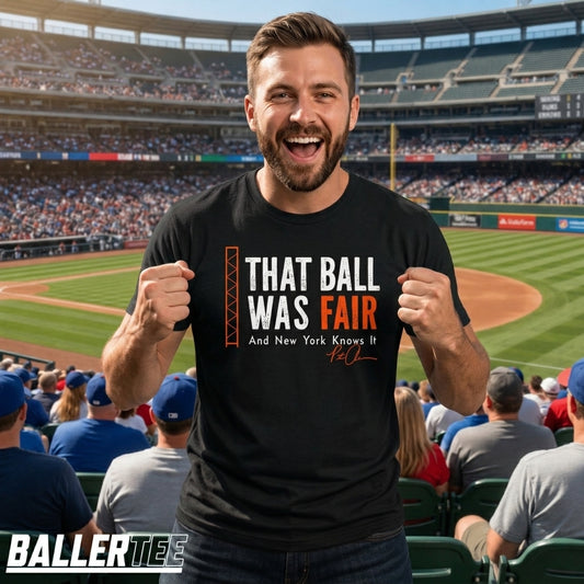 A man in a baseball stadium wearing a black graphic t-shirt that reads 'THAT BALL WAS FAIR And New York Knows It' with an orange and white design. The BallerTees logo is visible in the bottom-left corner.