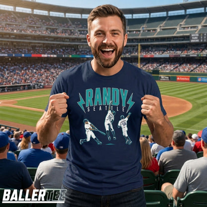 A man wearing a navy blue graphic t-shirt with a baseball player design and the text 'RANDY SEATTLE' stands in a crowded baseball stadium. The BallerTee logo is visible in the bottom-left corner.