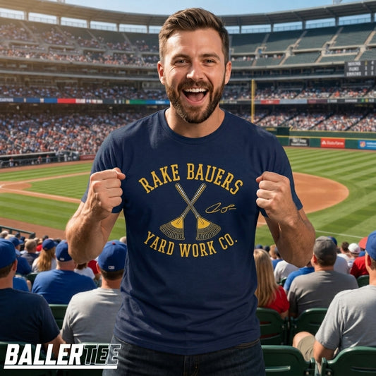A man in a baseball stadium wearing a navy blue t-shirt with a yellow graphic that reads 'RAKE BAUERS YARD WORK CO.' and features crossed golf clubs.