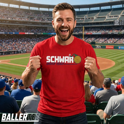 A man wearing a red graphic t-shirt with 'SCHWARZ since 1994' and a tennis ball graphic, standing in a crowded baseball stadium. The BallerTee logo is visible in the bottom-left corner.