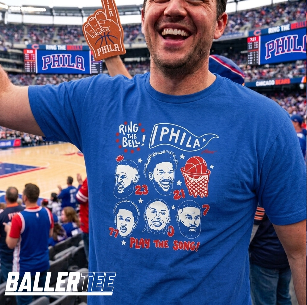 A man in a crowded basketball arena wearing a blue graphic T-shirt with Philadelphia Phillies-themed artwork and text, holding a foam finger. The shirt features illustrated faces, numbers, a basketball hoop, and the word 'PHILA'.