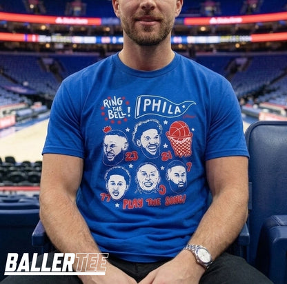 A man wearing a royal blue graphic t-shirt with a Philadelphia 76ers theme, seated in a stadium. The shirt features caricatures of basketball legends, the number 23, and the text 'RING THE BELL! PHILADELPHIA PLAY THE SONG!'.